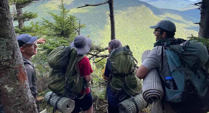 a group of people wearing backpacking gear stand among trees looking out over a green landscape far below